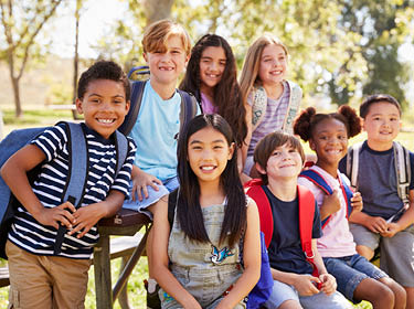 Students wear backpacks and sit at a picnic table in the shade of some trees