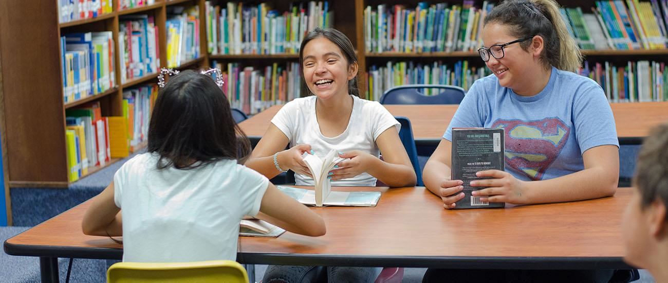 Photo of three students sitting at a table, holding books and talking